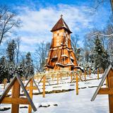 Cmentarz z drewnianymi nagrobkami, za nimi wysoka drewniana kaplica. Wszystko pokryte śniegiem.
