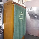 Duża gwiazda Dawida na zielonej tkaninie zasłaniającej szafę, na ścianach zdjęcia.