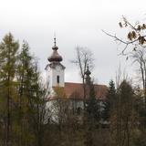 Kościół o prostej bryle z wieżą z cebulastym hełmem, biały z miedzianymi dachami. Widok jesienny z daleka, z boku zza bezlistnych drzew.