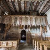 Widok na chór w drewnianym kościele w Dębnie z niedużymi organami, z sufitem malowanym w paski z różnymi wzorami z opadającą jakby koronką z balustrady z chóru. Z ławkami drewnianymi w dwóch rzędach, z widokiem na drzwi wejściowe z przejściem z łukiem oraz z otwartymi drzwiami bocznymi  przed ławkami po lewej. Na ścianach obrazy świętych i krzyż po prawej.