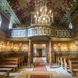 Organy w drewnianym kościele. Polichromowane ściany i stropy oraz barierki chóru muzycznego, kryształowy żyrandol, drewniane ławki.