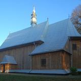 Kościół drewniany widziany z zewnątrz od strony prezbiterium. Oszalowane deskami ściany, blaszany dach, wkoło zieleń.
