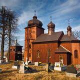 Kilka nagrobków na trawie. Za nimi drewniana cerkiew kryta blaszanym dachem z charakterystycznymi dla cerkwi trzema kopułami. Przy cerkwi po lewej stoi drewniana dzwonnica z małą kopułą na dachu. Wokół  kilka drzew bez liści. W tle po lewej łąki i wzgórza. Na niebie cienkie warstwy chmur.