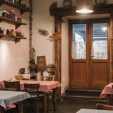 Wnętrze restauracji. Na wprost znajdują się drzwi, pod ścianami stoliki przykryte obrusami w kratkę, na półkach słoiki z przetworami.