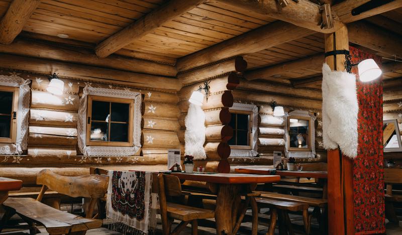 Wnętrze karczmy z drewnianych bali. Drewniane stoły i ławy, białe zdobienia wokół okien.