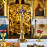 Metalowy krzyż i kielichy na wąskim ołtarzu w drewnianej cerkwi. Przed nimi leży księga i drewniany krzyż. W tle rozmyty widok na bogate carskie wrota i obok obrazy świętych.
