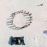 Snowboardziści podczas rozgrzewki w kółku uchwyceni z drona na białym stoku narciarskim Podstolice Ski. Obok nich stoi ratrak.