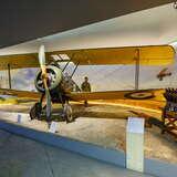 Mały samolot wojskowy w hangarze w Muzeum Lotnictwa Polskiego w Krakowie. Obok niego stoją eksponaty wojenne, a z tyłu postać z kartonu.